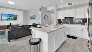 A modern kitchen with white cabinets, a stainless steel refrigerator, and a dining area with a gray sofa and a flat-screen TV. - Storey Lake Resort 2 Bedroom Condo with Private Balcony 2 Condo (Kissimmee)