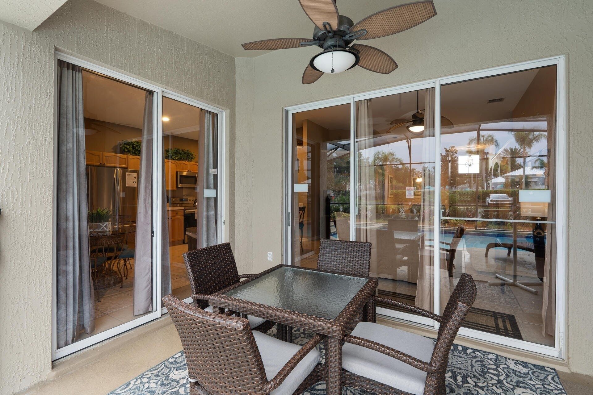 Un espace patio avec une table en verre et des chaises en osier, un ventilateur de plafond, et une vue sur une piscine et du mobilier d’extérieur.