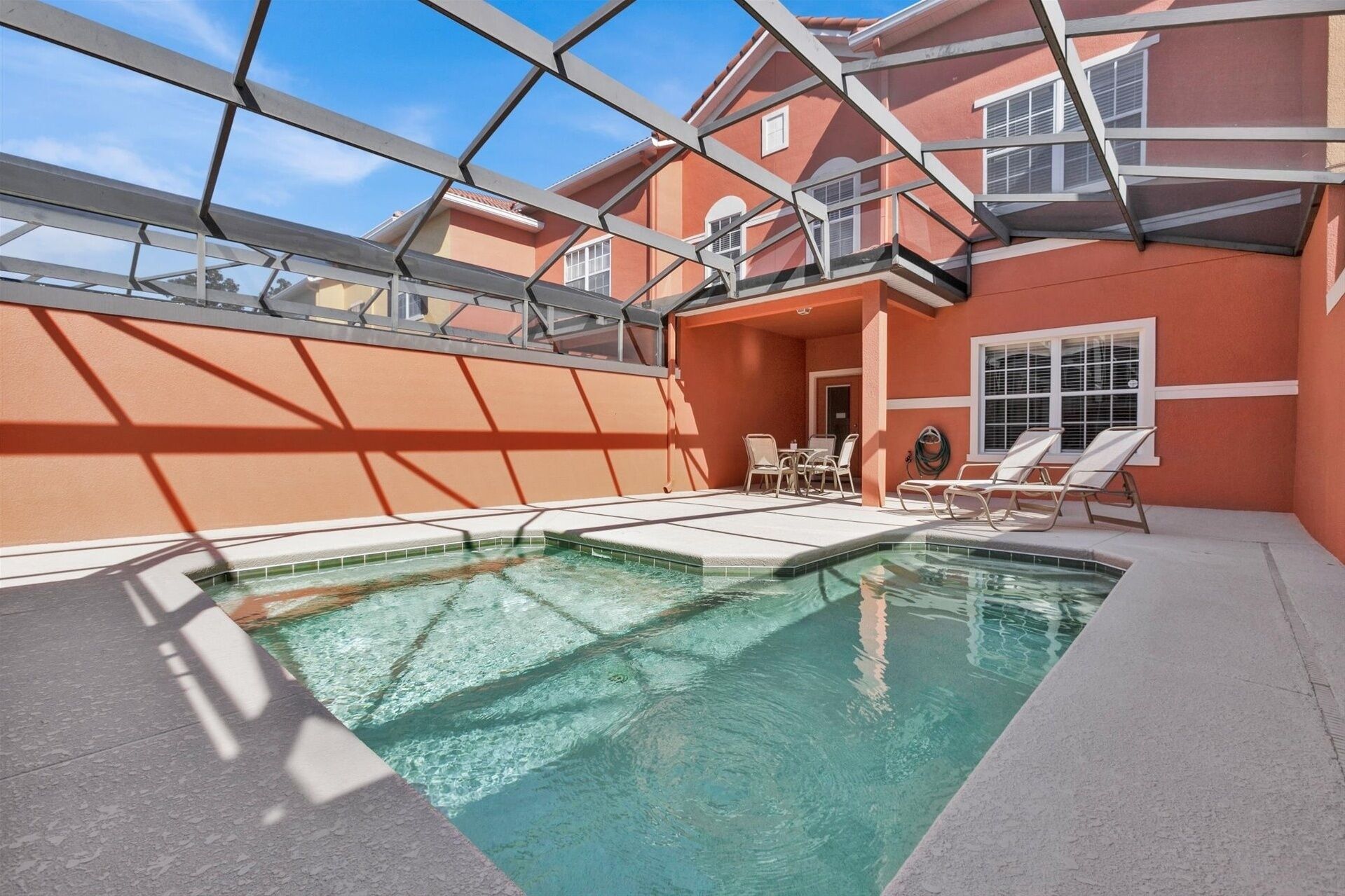 An indoor swimming pool with a glass ceiling, surrounded by a red building with a patio area and lounge chairs.