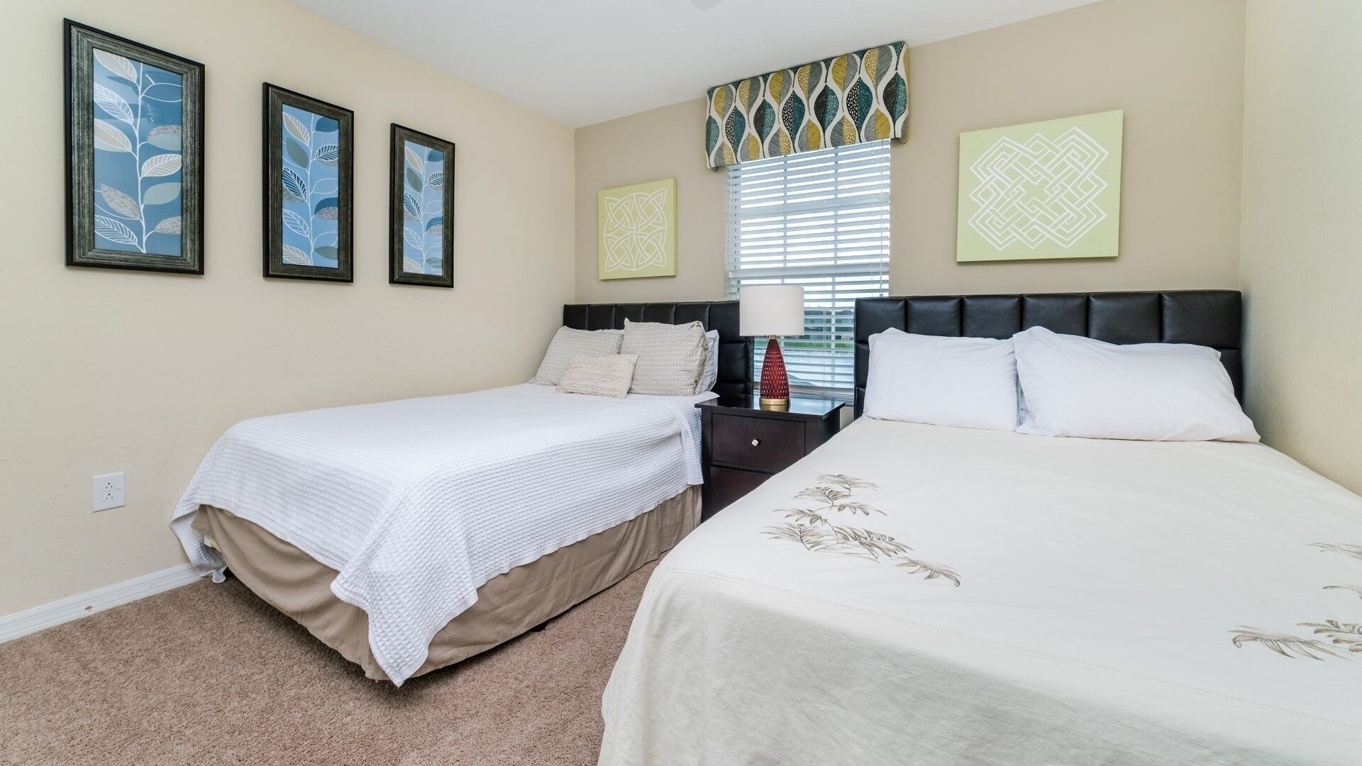 A hotel room with two beds, a nightstand, a lamp, and three framed artworks on the wall.