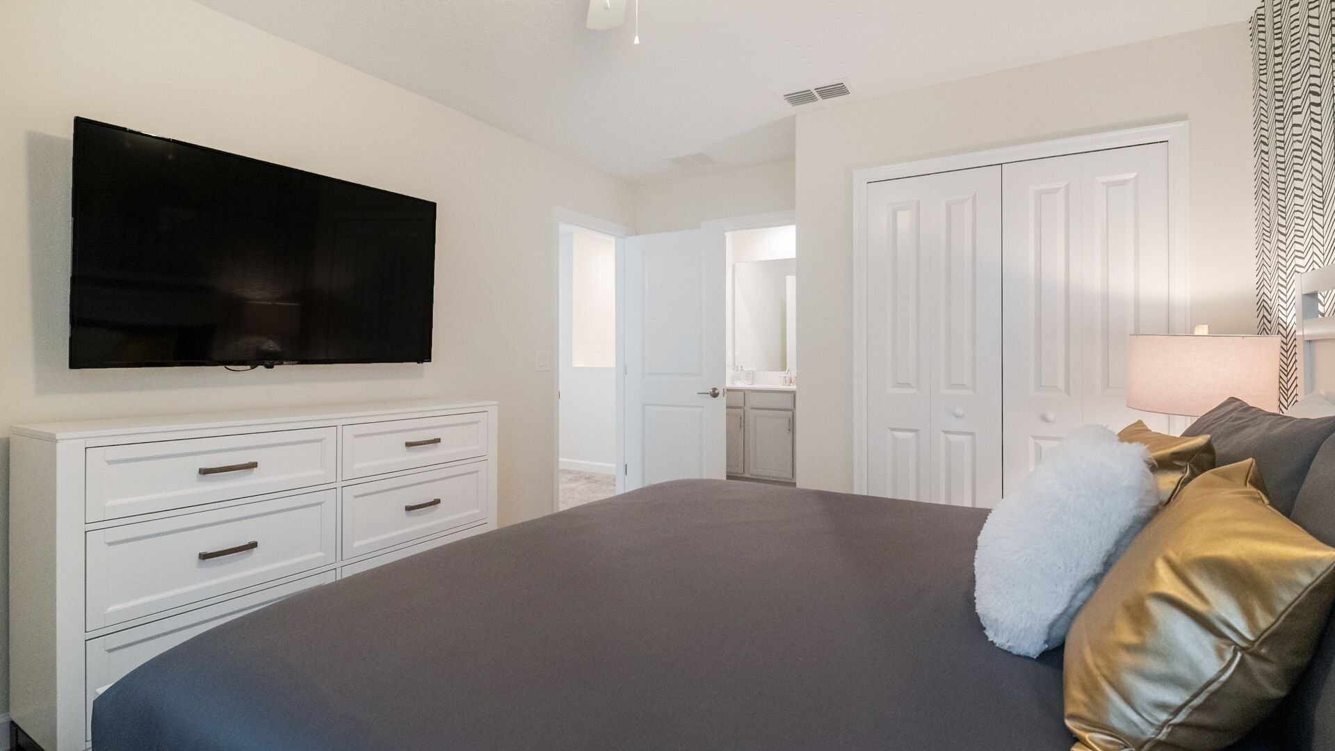 A bedroom with a large bed, a TV mounted on the wall, a dresser, and a door leading to another room.