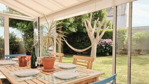 Une table en bois, dressée pour le repas, avec des chaises, une plante en pot et un hamac, le tout donnant sur un jardin.