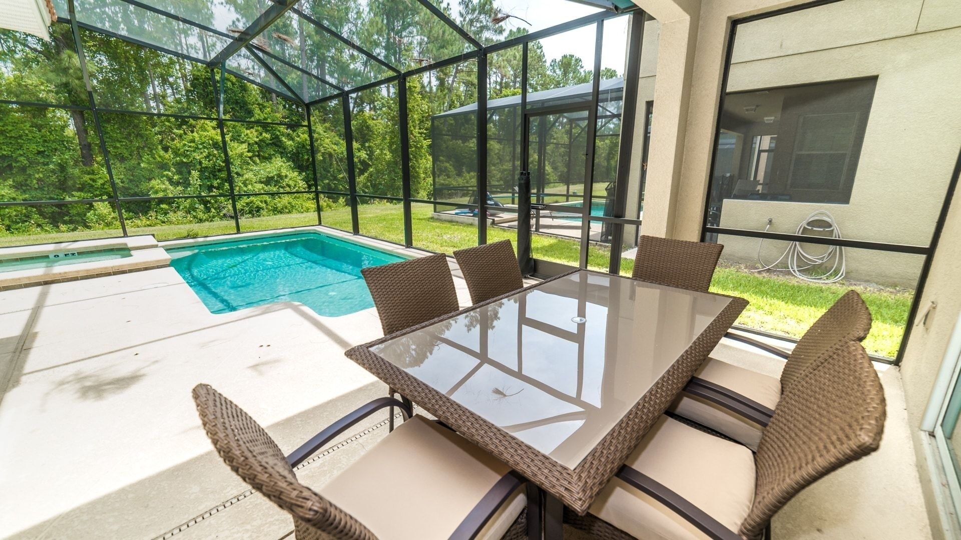 An outdoor patio with a glass table and wicker chairs, overlooking a swimming pool and lush greenery.