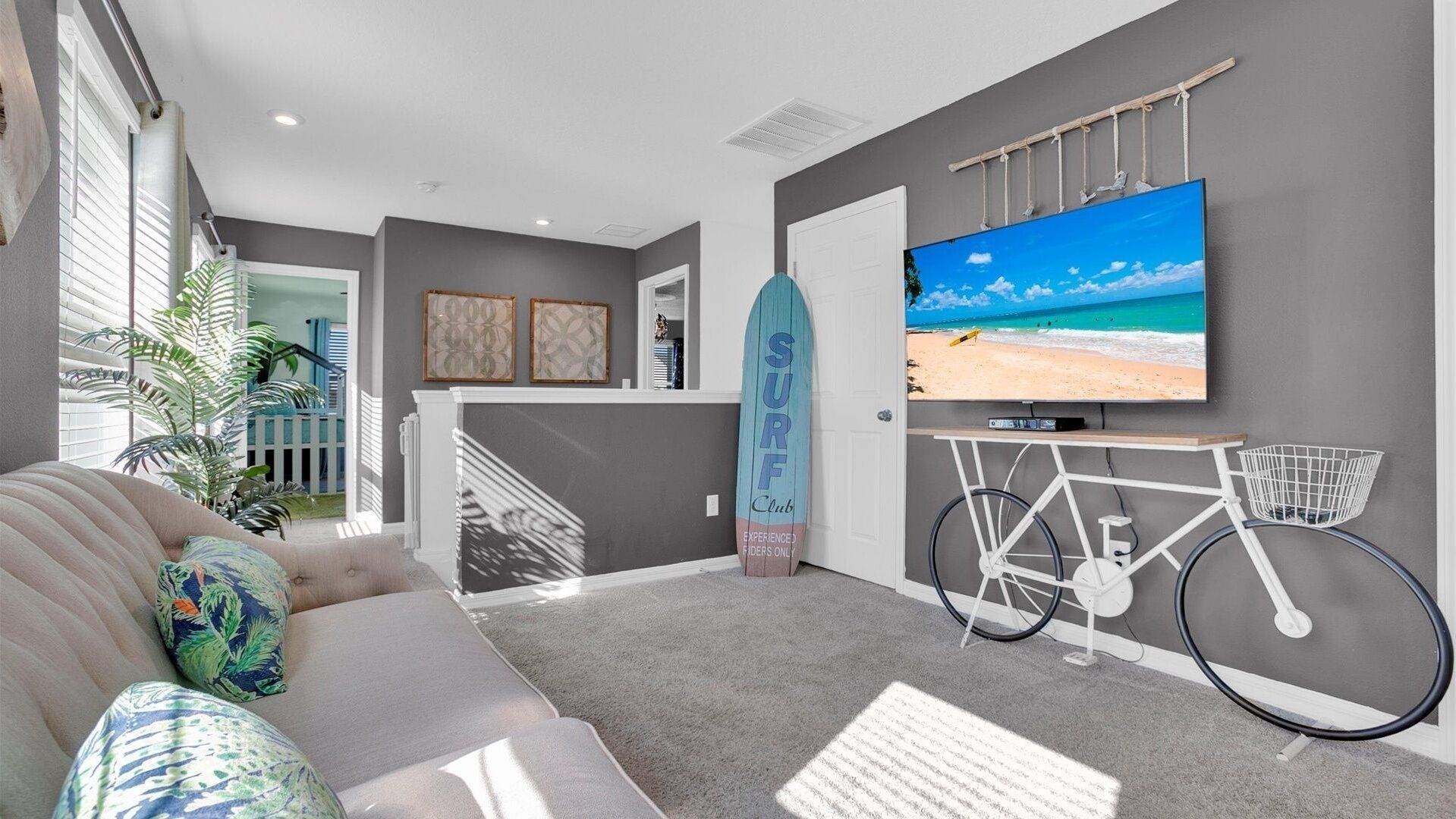 Una sala de estar moderna con un sofá gris, una tabla de surf, una bicicleta y un televisor mostrando una escena de playa.