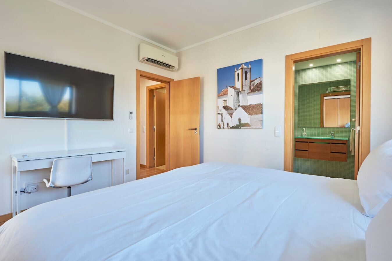 Une chambre d’hôtel avec un grand lit, un bureau avec une chaise, une télévision, un tableau et une salle de bain dont un mur est recouvert de carreaux verts.