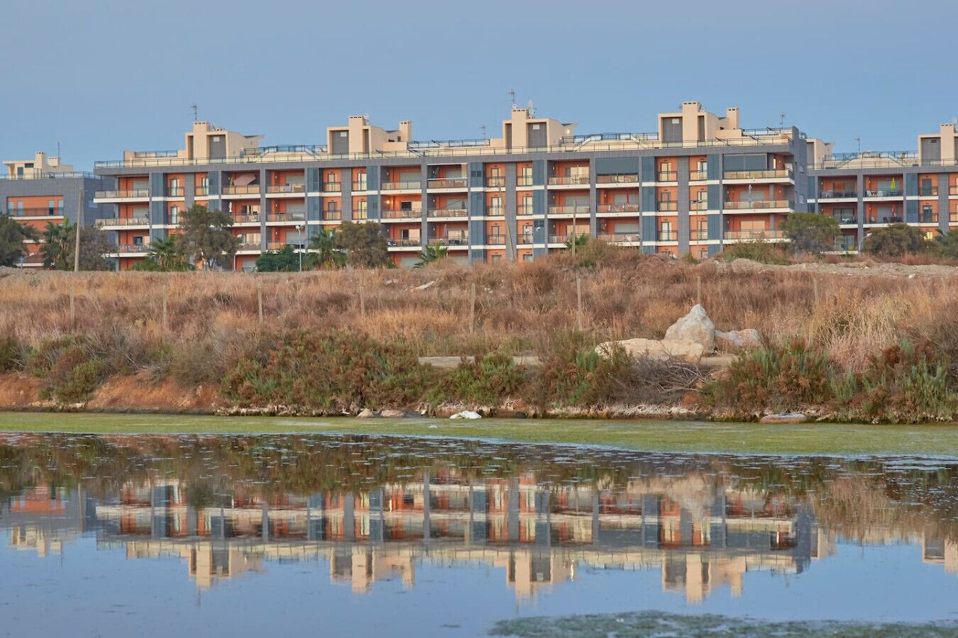 Un immeuble résidentiel à plusieurs étages, doté de balcons, situé au bord d’un plan d’eau dans lequel se reflète le bâtiment.