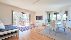 Een moderne woonkamer met een bank, een televisie, een eethoek met stoelen en grote ramen met gordijnen.