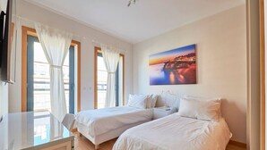 Een hotelkamer met twee bedden, een bureau en een schilderij van een zonsondergang boven een kustlandschap.