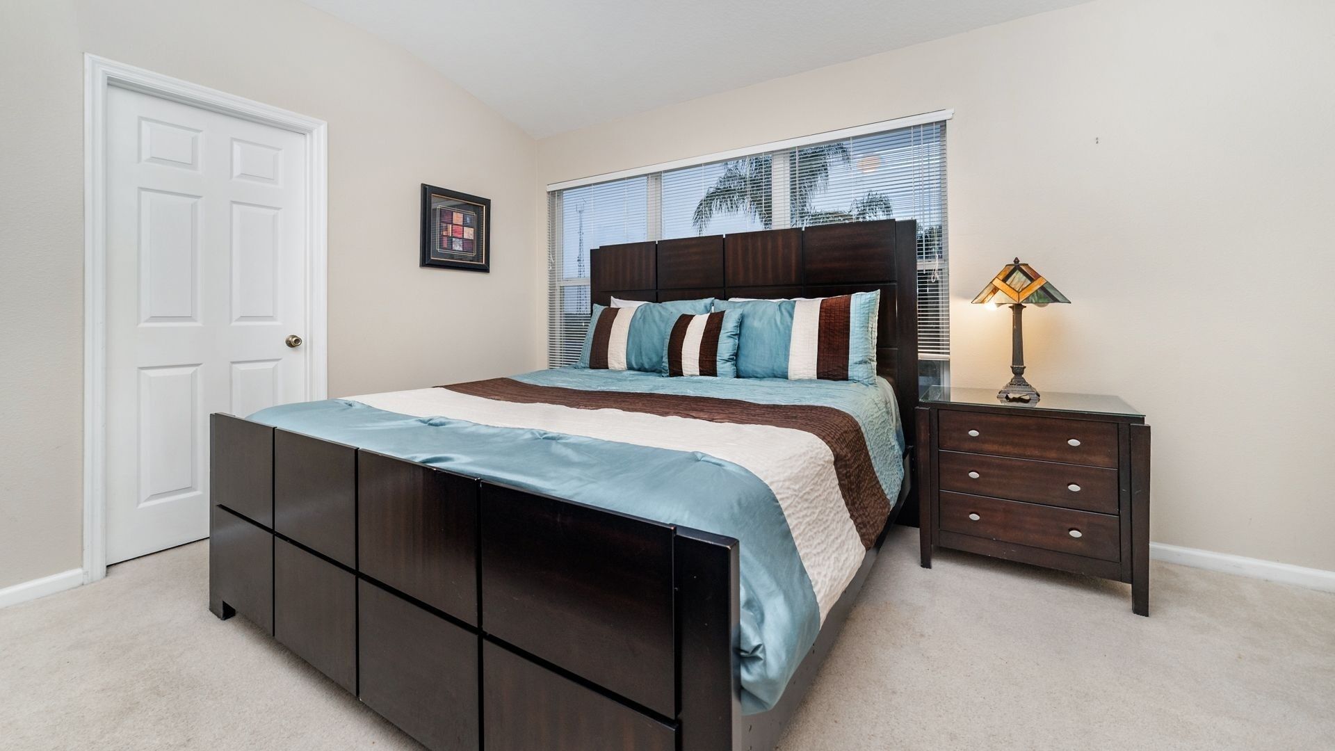 A bedroom with a bed, a nightstand, a lamp, a door, and a window with blinds.