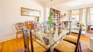Un comedor con mesa de vidrio, sillas de madera, lámpara de araña, estantería y sofá en una habitación luminosa con ventanas amplias.