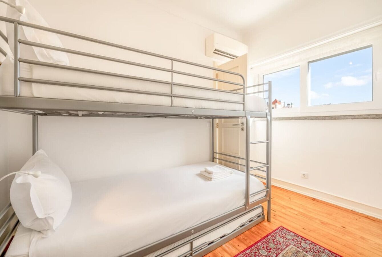 Uma cama beliche com colchão e travesseiro brancos, uma escada de metal e um tapete vermelho estampado sobre piso de madeira.