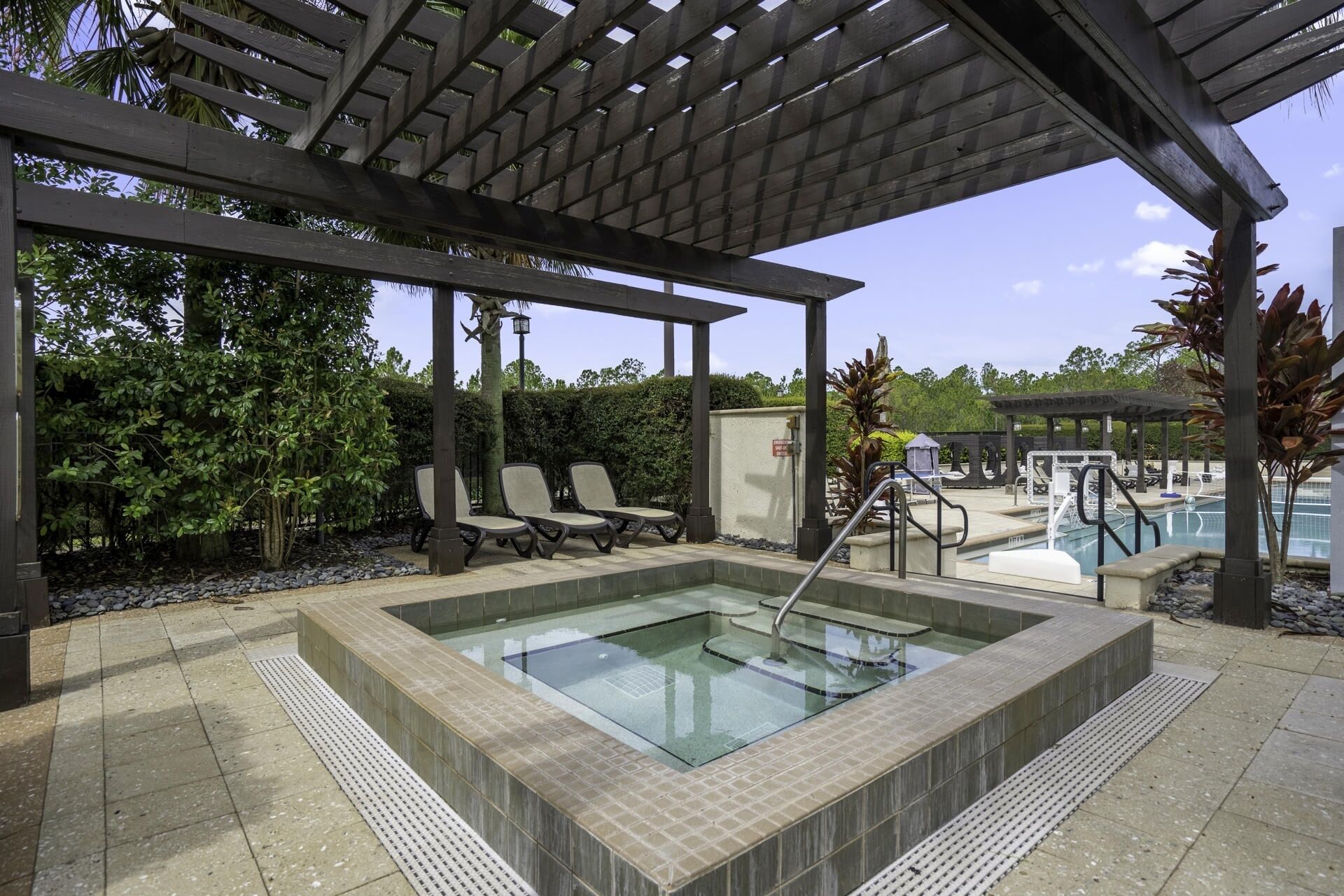 Un espace avec un jacuzzi, surmonté d’une pergola, entouré de chaises longues et d’une piscine.