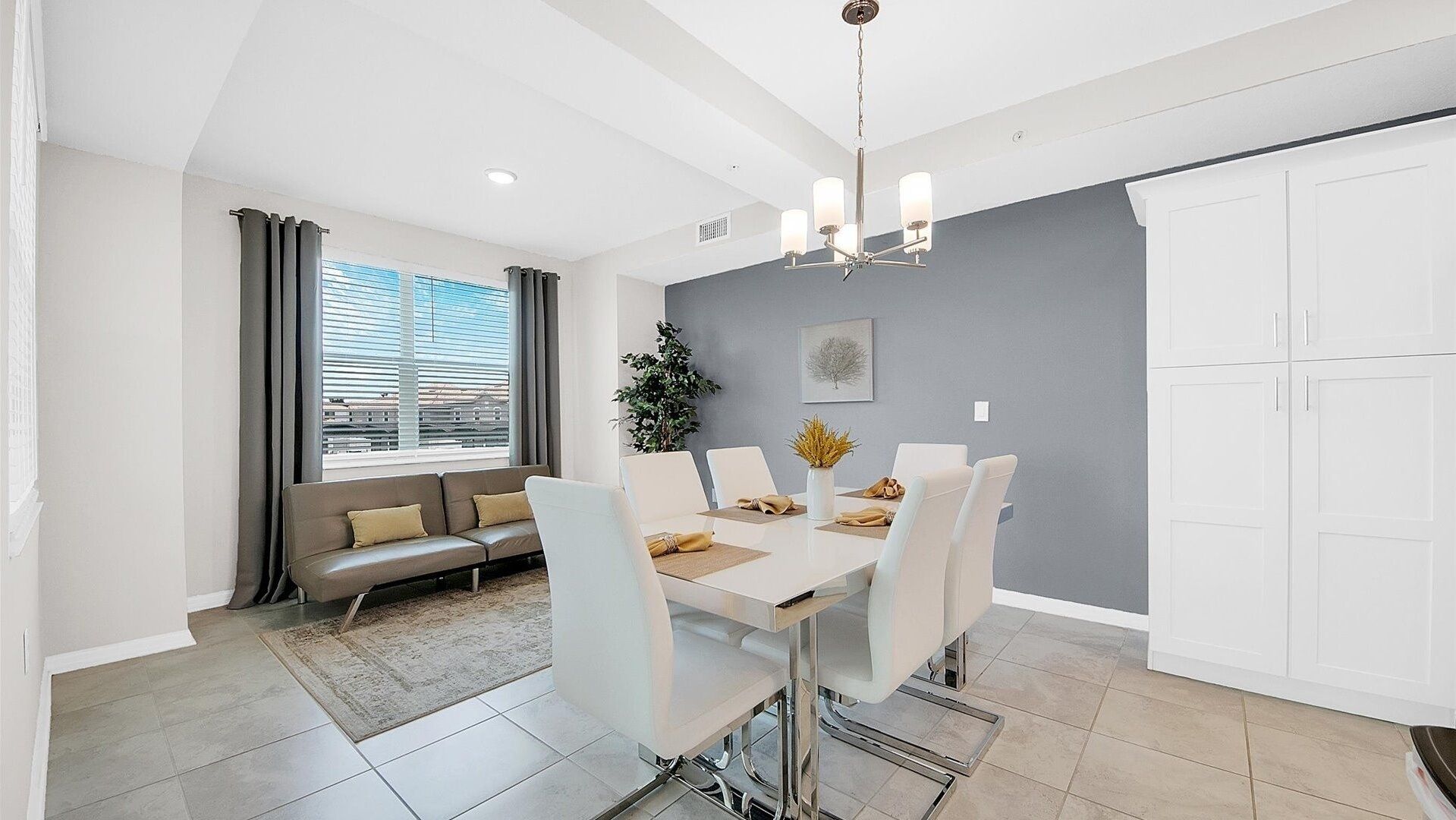 Un espace de salle à manger moderne avec une table blanche, des chaises et un lustre. Un mur gris est orné d’un tableau encadré, et une fenêtre est dotée de stores.