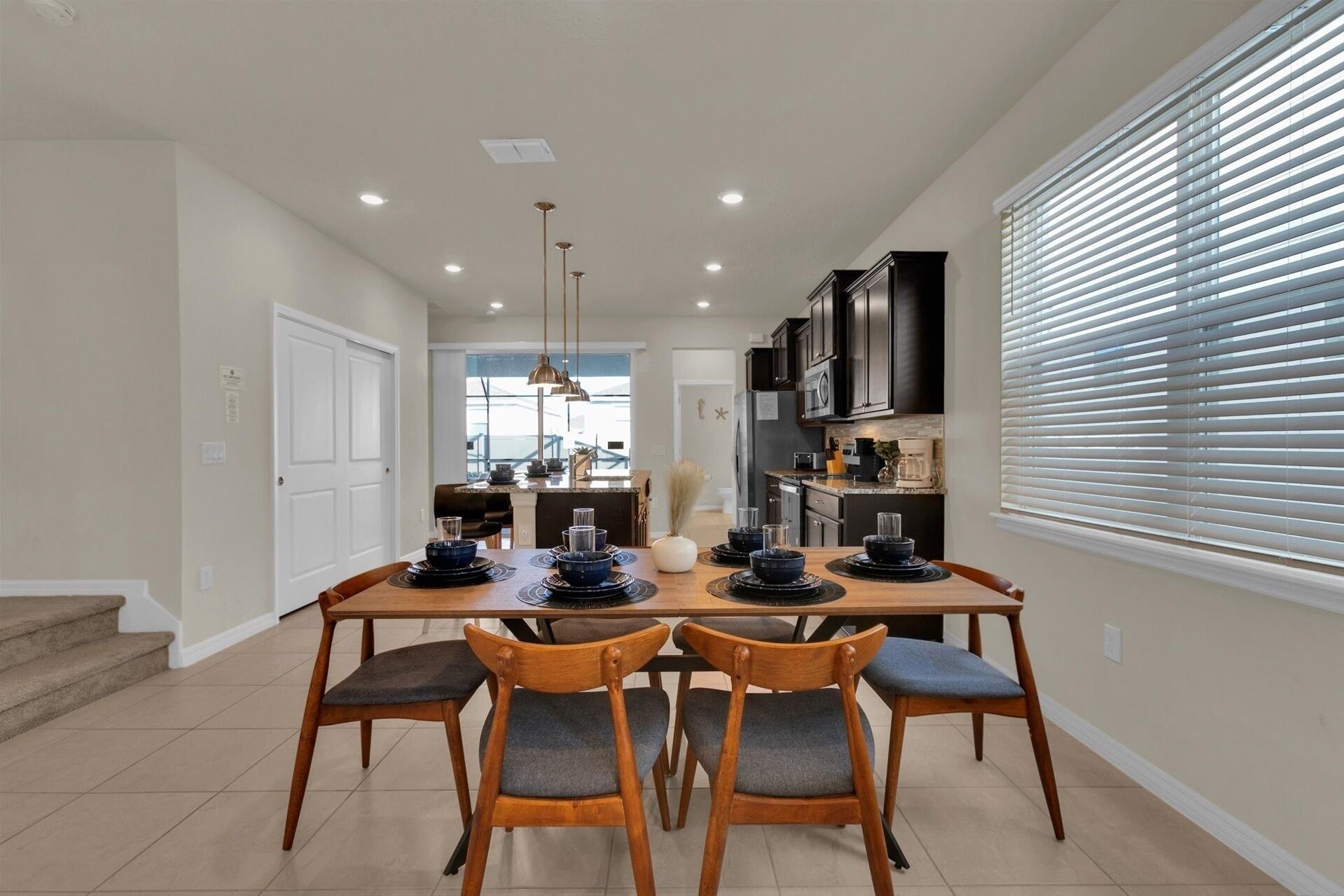 A dining area with a wooden table and chairs, a kitchen in the background, and a window with blinds.