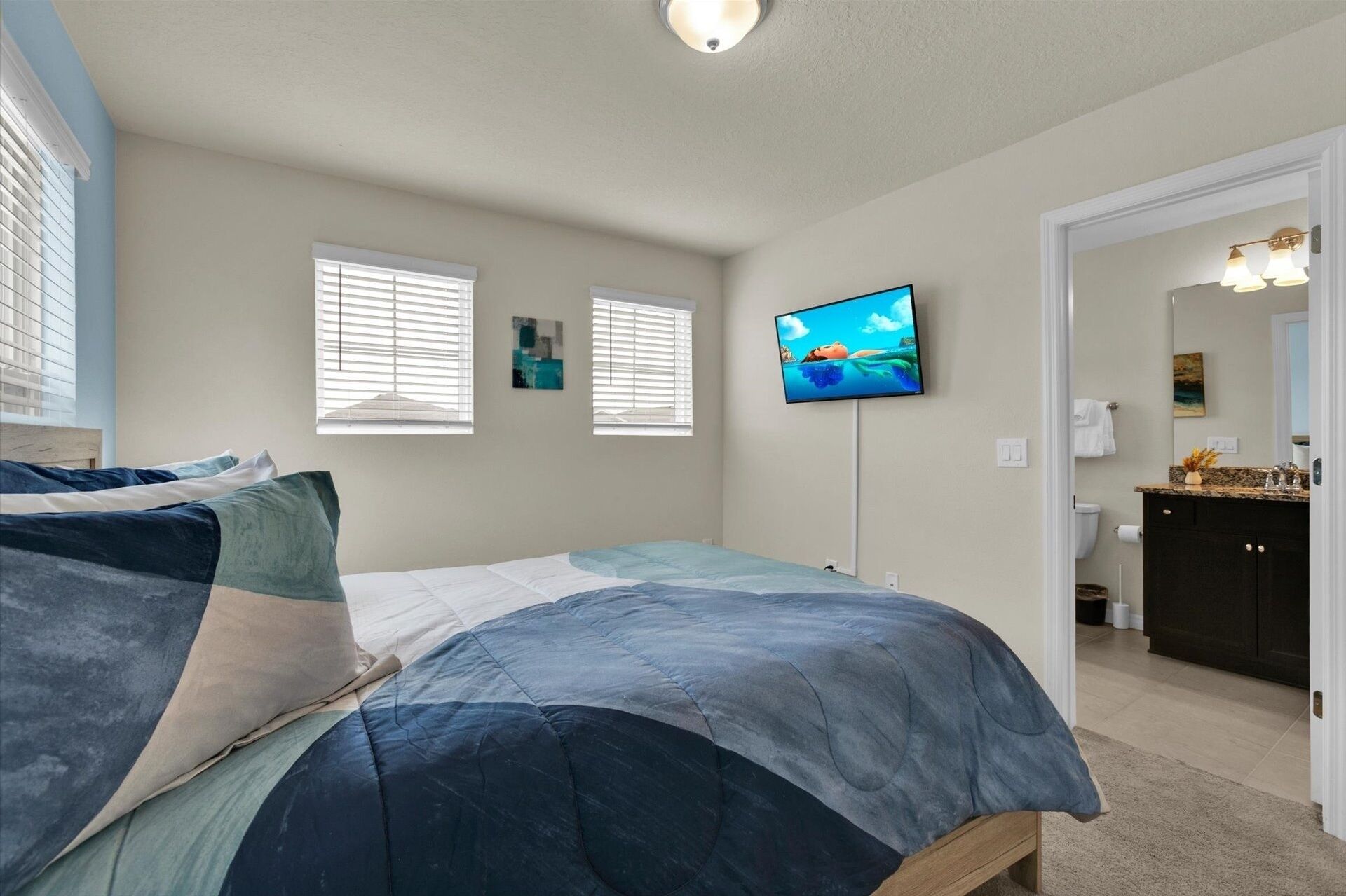 Un dormitorio con una cama, dos ventanas, un televisor y un baño visible a través de una puerta abierta.