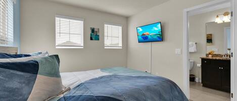 Un dormitorio con una cama, dos ventanas, un televisor y un baño visible a través de una puerta abierta.