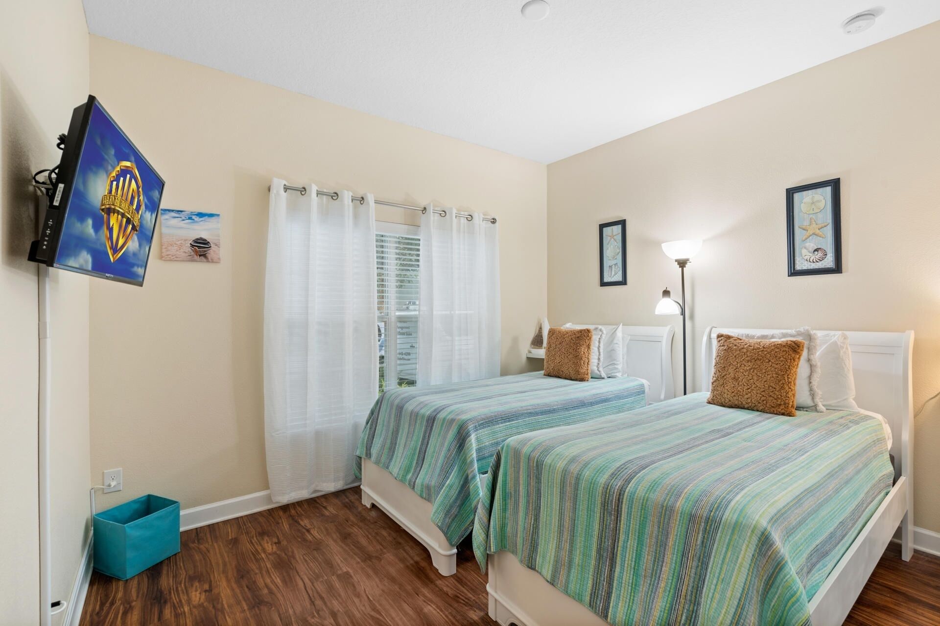 A bedroom with two beds, a TV mounted on the wall, a window with curtains, and a bedside lamp.