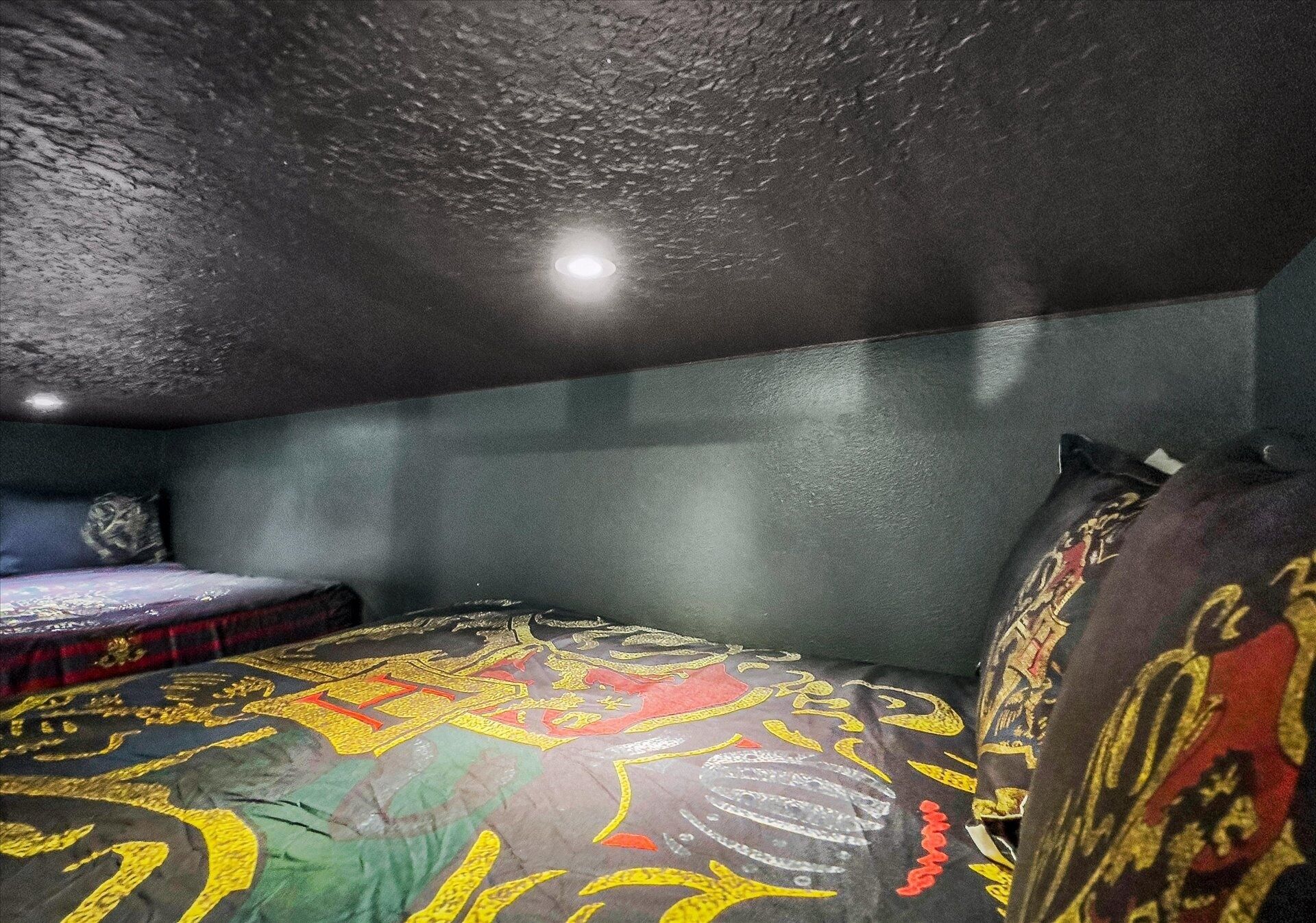 A room with a textured ceiling, a bed with a colorful patterned bedspread, and a small table with a decorative item.