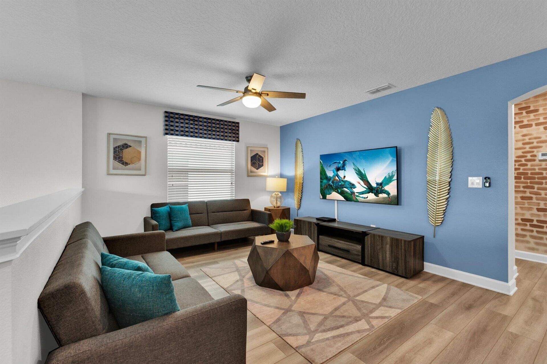 A modern living room with a blue accent wall, a ceiling fan, a large flat-screen TV, and a brick accent wall.