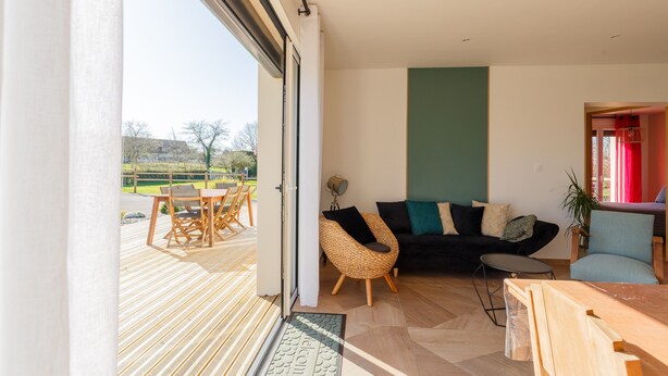 A modern living room with a sofa, chairs, a dining table, and a view through a large sliding door to an outdoor deck. - Gîte 'Le Nid Des Prés' with Private Terrace and Garden (Val-de-Scie)
