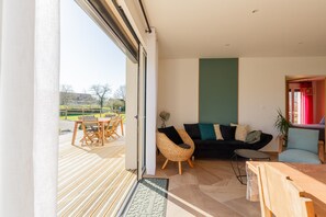 A modern living room with a sofa, chairs, a dining table, and a view through a large sliding door to an outdoor deck. - Gîte 'Le Nid Des Prés' with Private Terrace and Garden (Val-de-Scie)