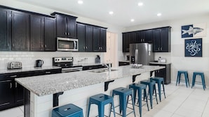 Eine moderne Küche mit dunklen Schränken, einer Arbeitsplatte aus Granit, einem Edelstahlkühlschrank, einer Mikrowelle, einem Toaster und blauen Barhocker.
