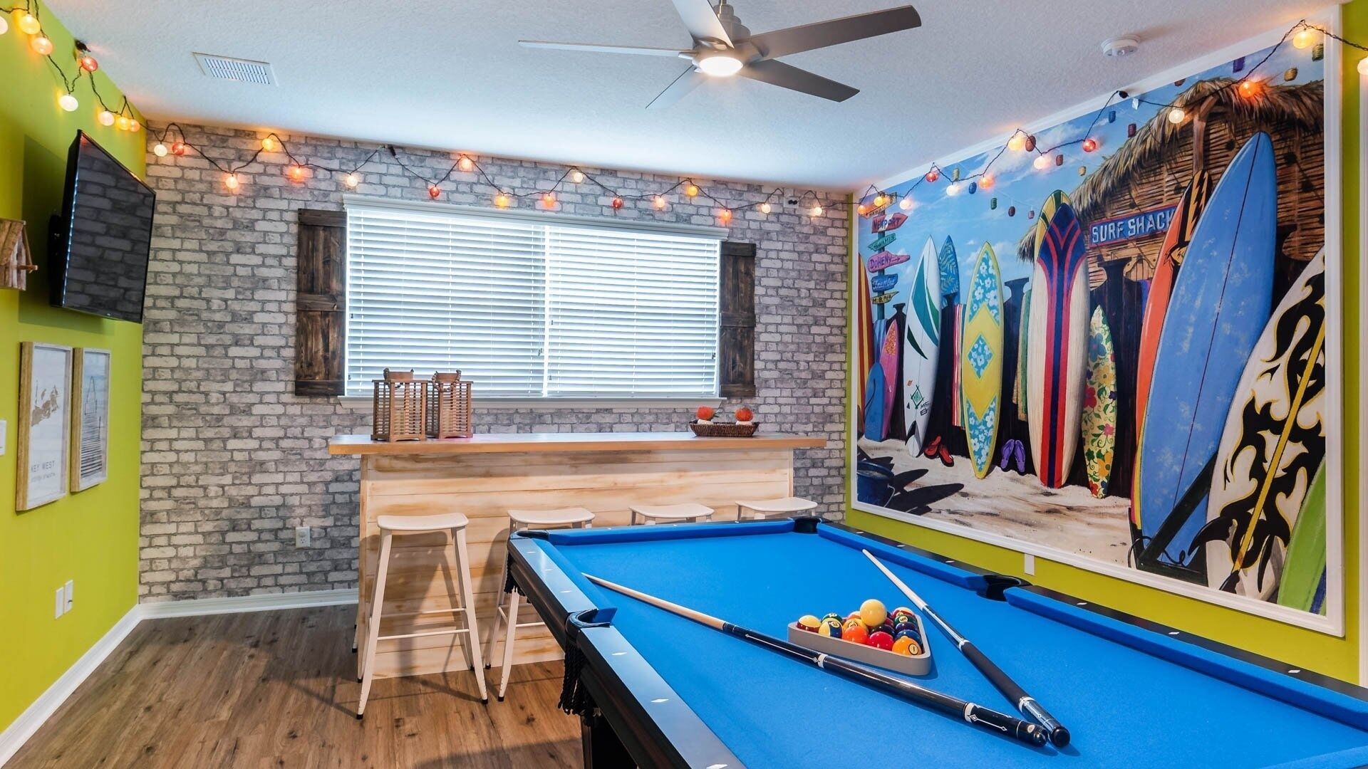 Une table de billard avec des queues et des billes, un bar avec des tabourets, une fresque représentant des planches de surf et un ventilateur au plafond.