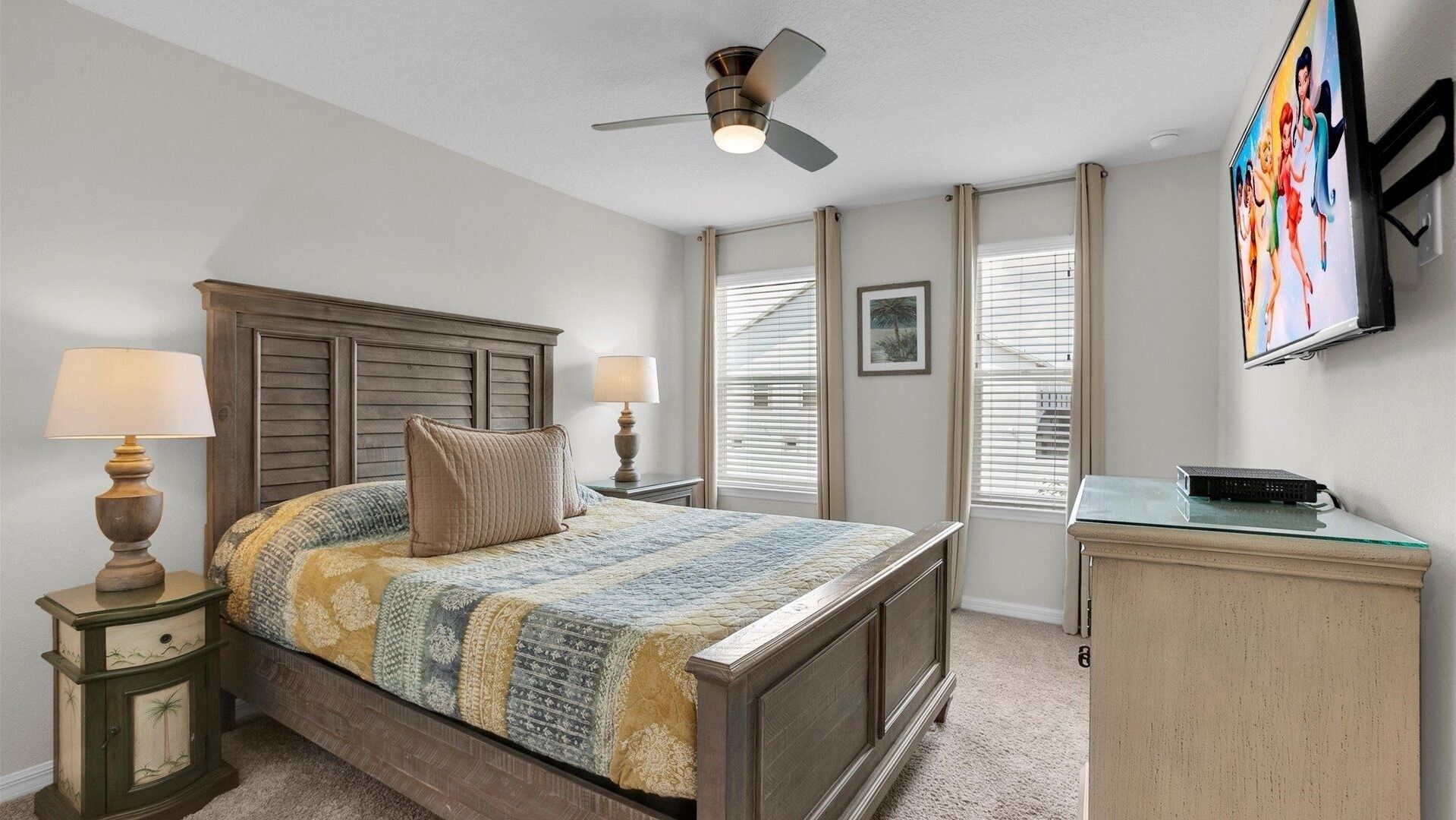 A bedroom with a wooden bed, two bedside lamps, a ceiling fan, a TV on a stand, and a window with blinds.