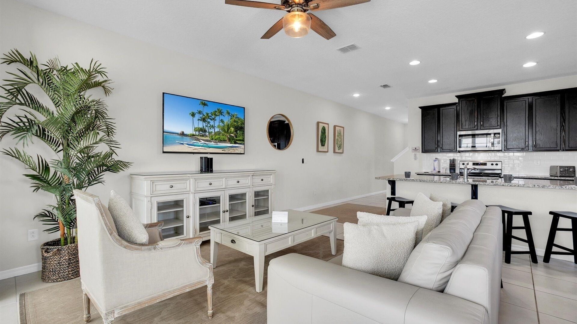 A modern living room with a white sofa, a glass coffee table, and a white cabinet. There is a large plant, a painting of a beach, and a ceiling fan.