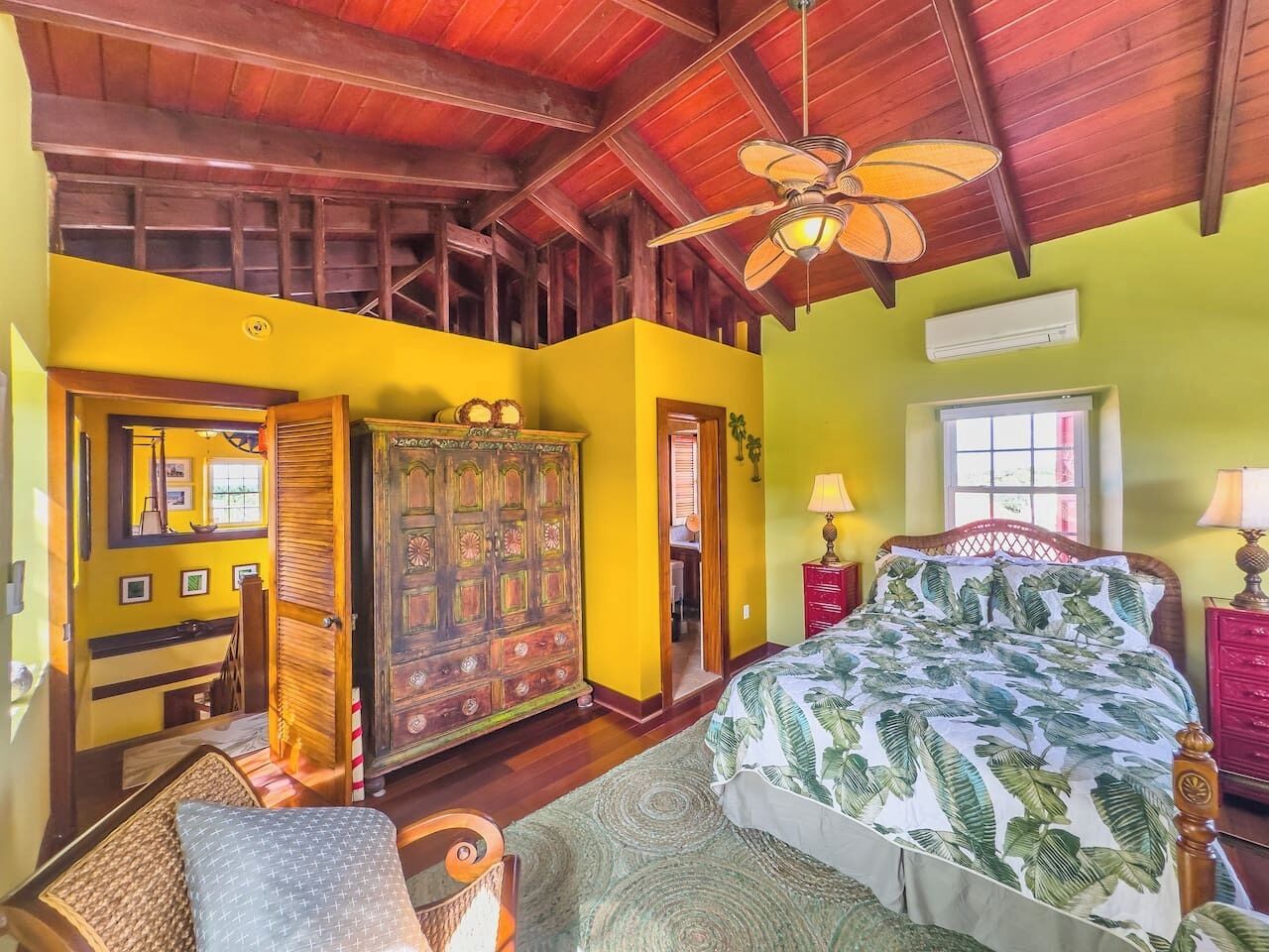 A bedroom with a wooden ceiling, a bed with a tropical print bedspread, a wooden wardrobe, and a red nightstand with a lamp.