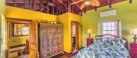 A bedroom with a wooden ceiling, a bed with a tropical print bedspread, a wooden wardrobe, and a red nightstand with a lamp.