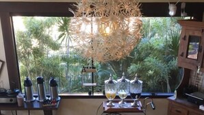 Un bufé de desayuno con diversas opciones de comida y bebidas, una lámpara colgante decorativa y vistas a follaje a través de la ventana.
