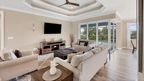 A spacious living room with a large sofa, a coffee table, and a TV. There is a ceiling fan and a large window offering a view of the outdoors. - Englewood 5 Bedroom Pool Home 3 miles to Gulf Beaches! 5 Home (Englewood)