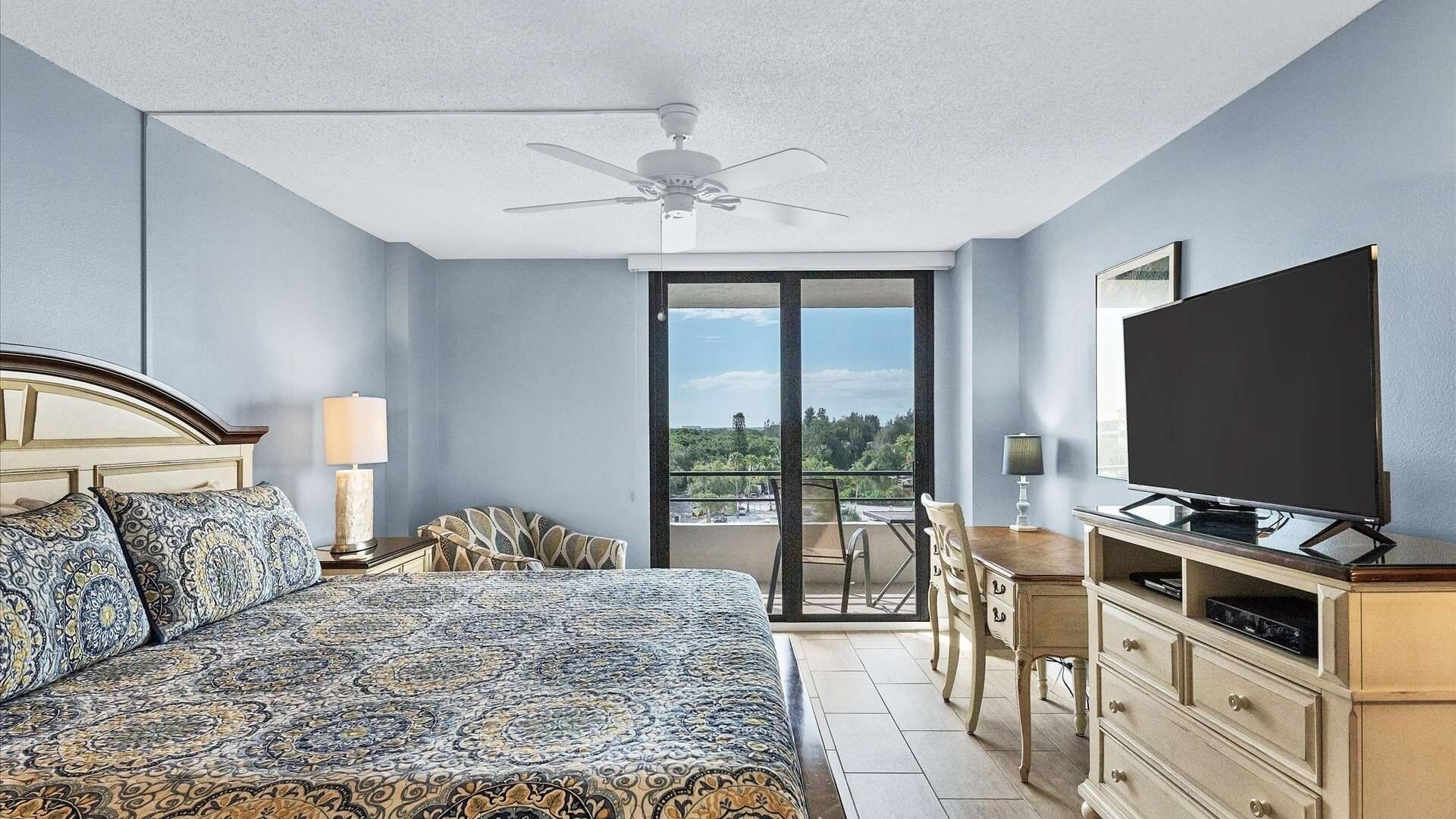 Quarto de hotel com uma cama grande, ventilador de teto, televisão, escrivaninha com cadeira e varanda com vista.