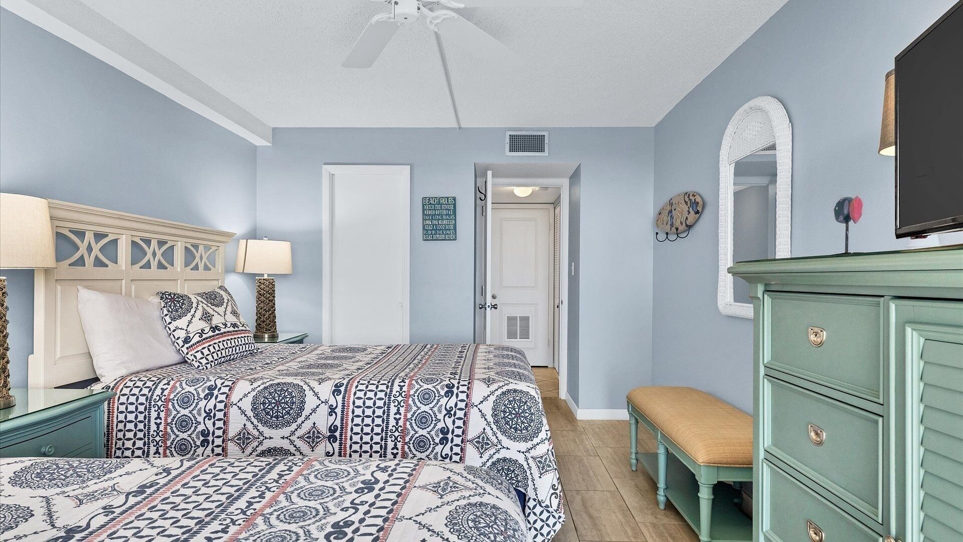 A bedroom with a patterned bedspread, a green dresser, a bench, and a ceiling fan.