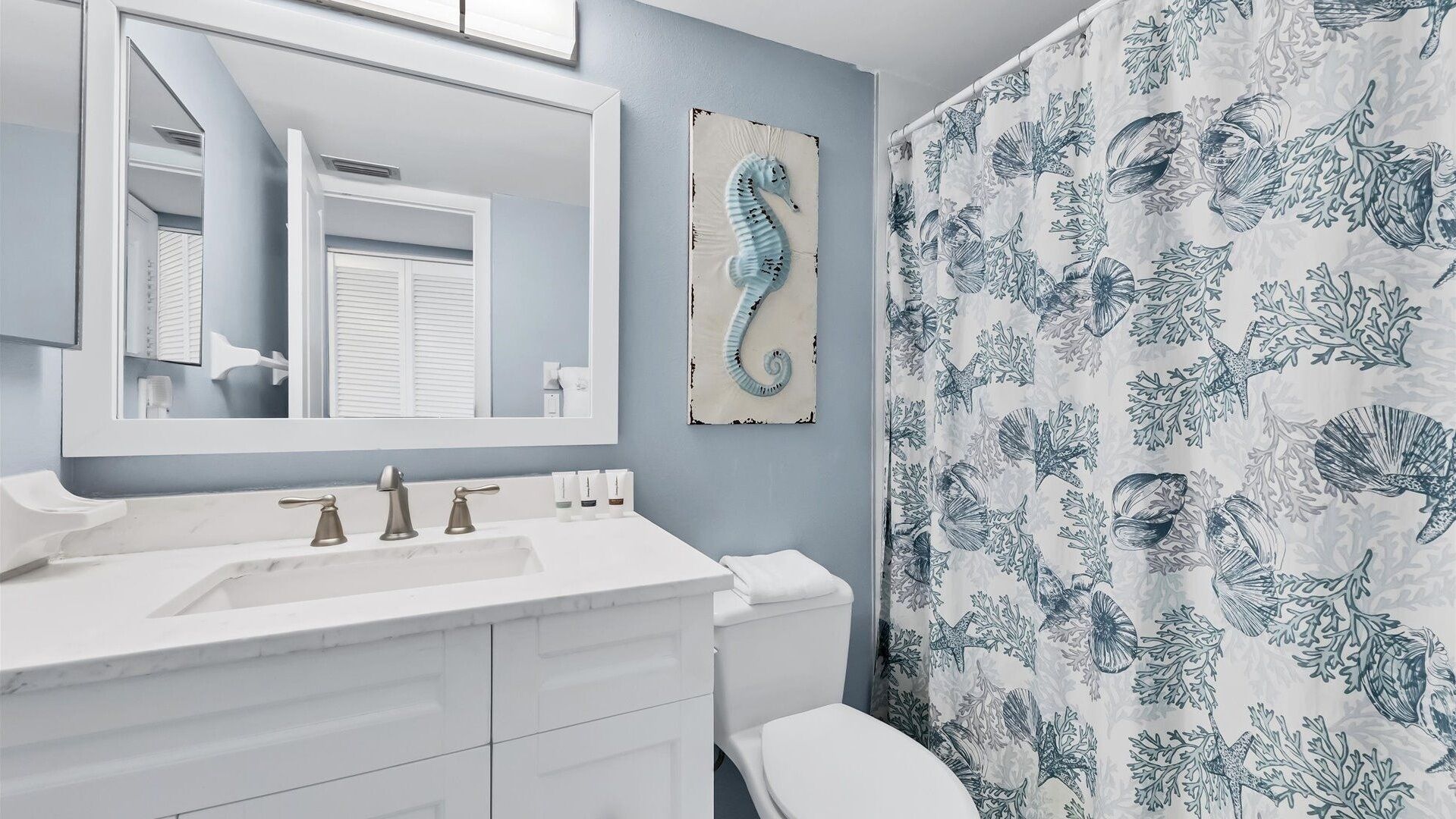 Une salle de bain avec un lavabo blanc, un miroir, une œuvre d’art représentant un hippocampe et un rideau de douche orné de motifs marins.
