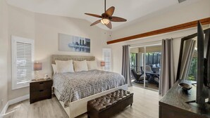Ein Schlafzimmer mit Bett, Nachttischen, Deckenventilator, Fenster mit Jalousien und Blick auf eine Außenpatio.