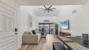 A modern living room with a sofa, a dining table with benches, a ceiling fan, and a TV on the wall. - Venice Beach Two Bedroom Private Condo with Community Pool, Gulf Beach Access and Water Views! 2 Condo (Venice)