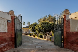 Uma entrada com portão, muro de tijolos, palmeira e um edifício com telhado amarelo ao fundo.