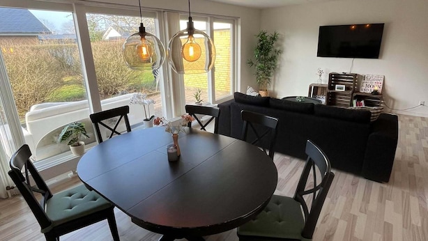 Un coin repas avec une table ronde et des chaises, un téléviseur fixé au mur et une vue sur l’extérieur grâce à de grandes fenêtres.