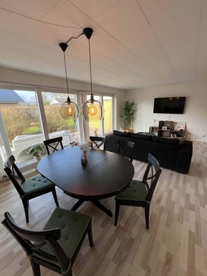 A dining area with a round table and chairs, a TV mounted on the wall, and a view of the outdoors through large windows.