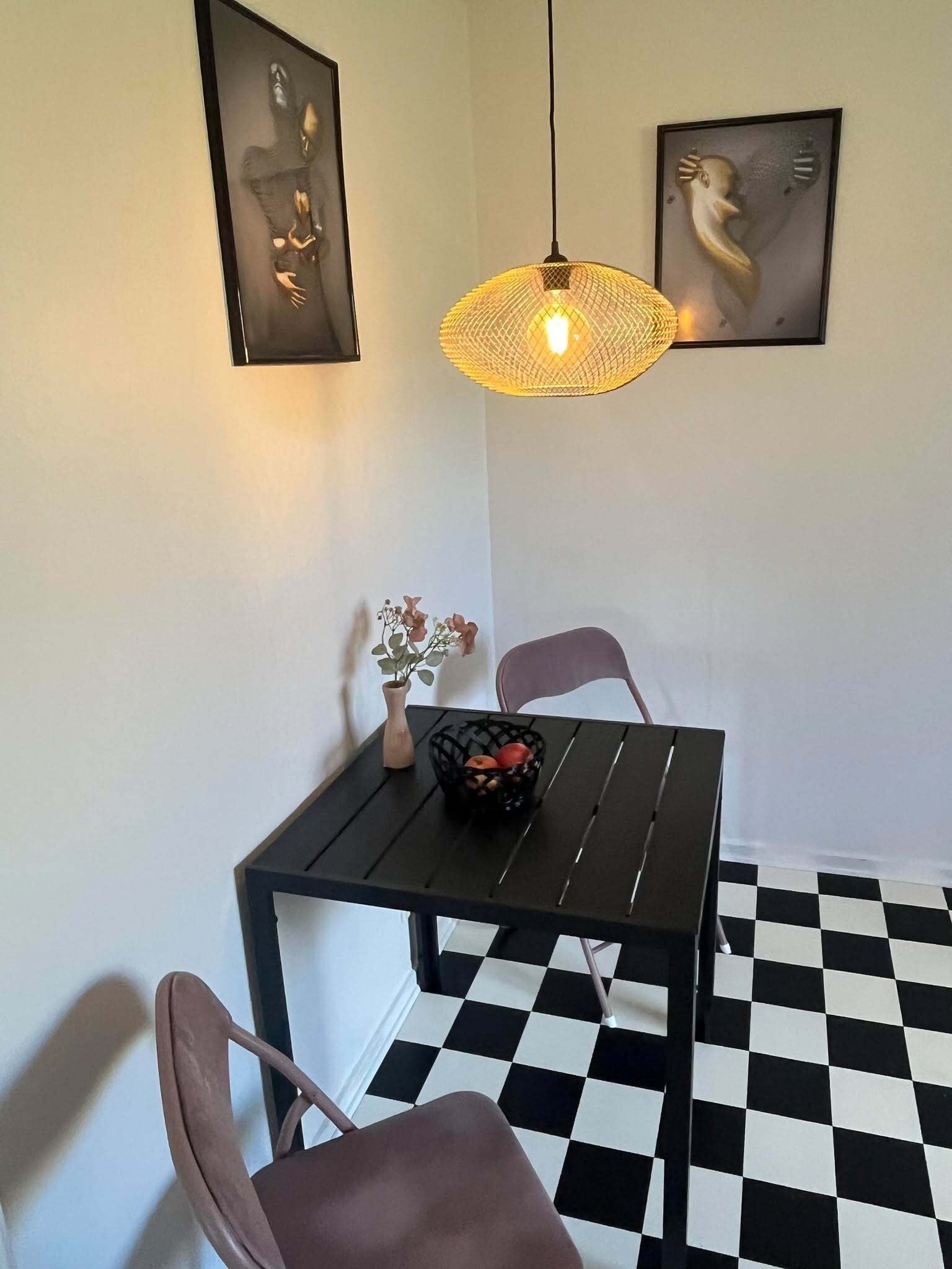 Un coin repas avec un sol à carreaux noirs et blancs, une table noire sur laquelle se trouve un panier de fruits, un vase rempli de fleurs et deux chaises.