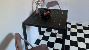 Un coin repas avec un sol à carreaux noirs et blancs, une table noire sur laquelle se trouve un panier de fruits, un vase rempli de fleurs et deux chaises.