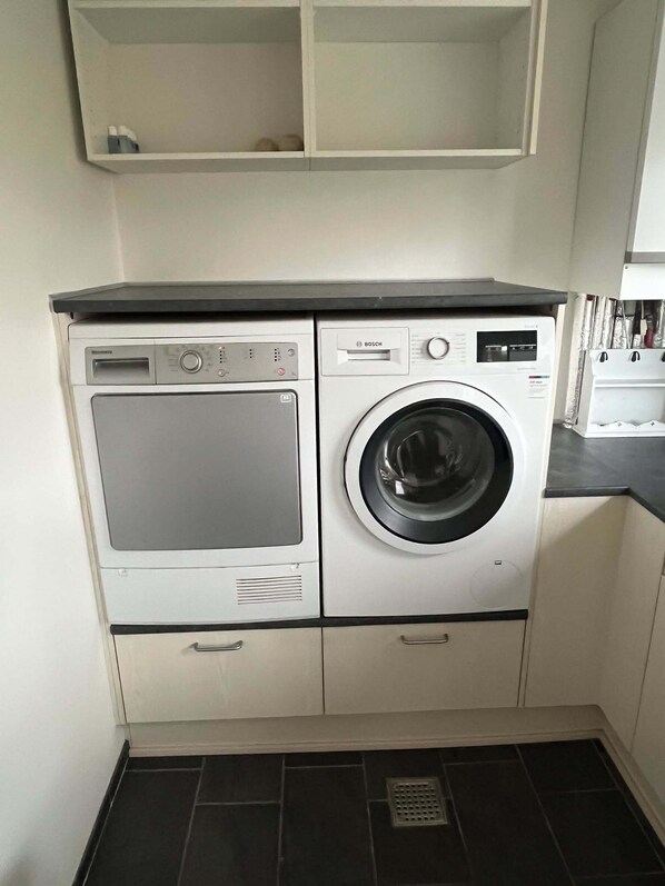 Une petite buanderie équipée d’un lave-linge et d’un sèche-linge à chargement frontal, de placards intégrés et d’un sol carrelé.