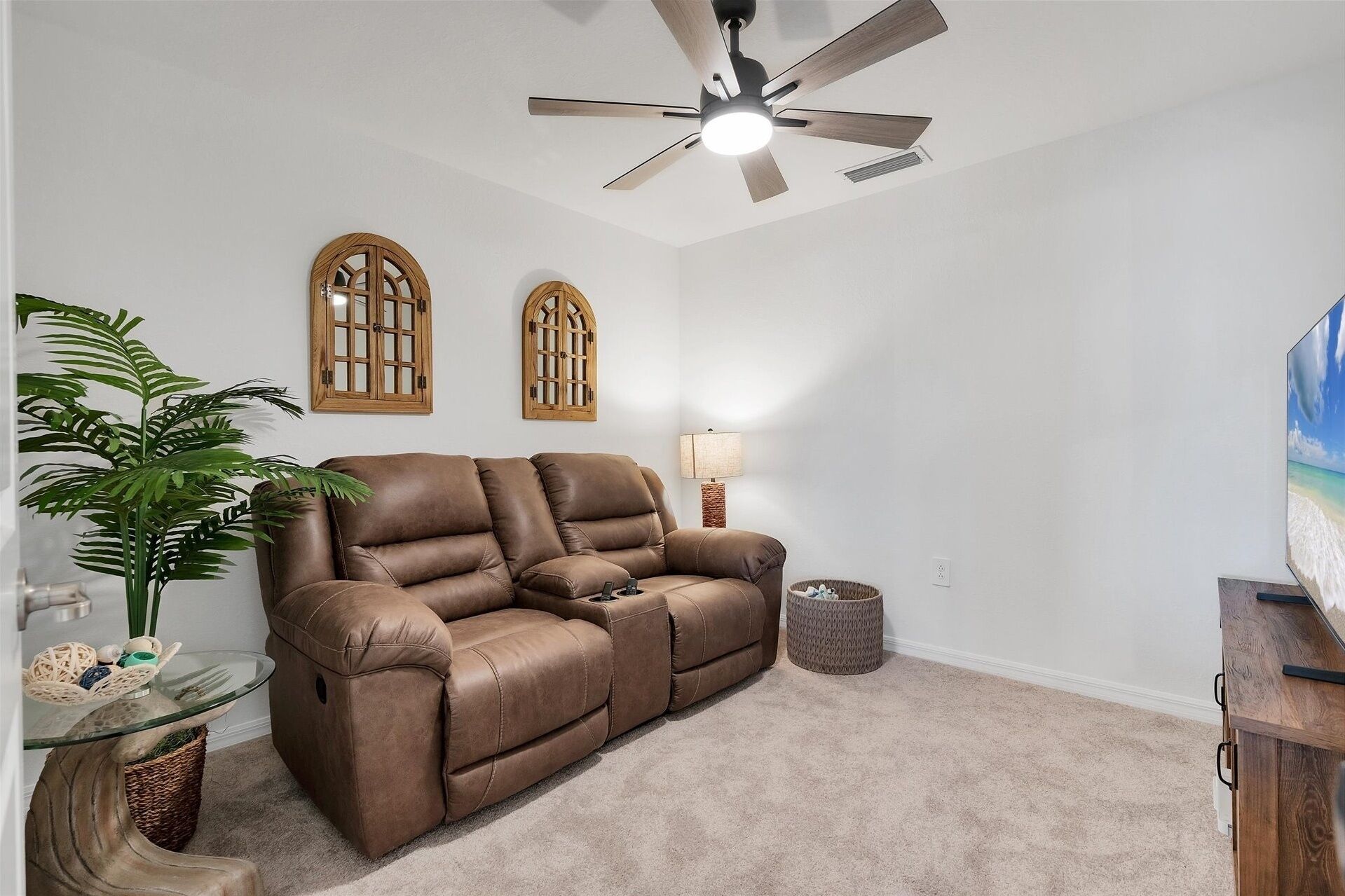 A living room with a brown leather sofa, a ceiling fan, two wooden-framed windows, a lamp, and a plant.