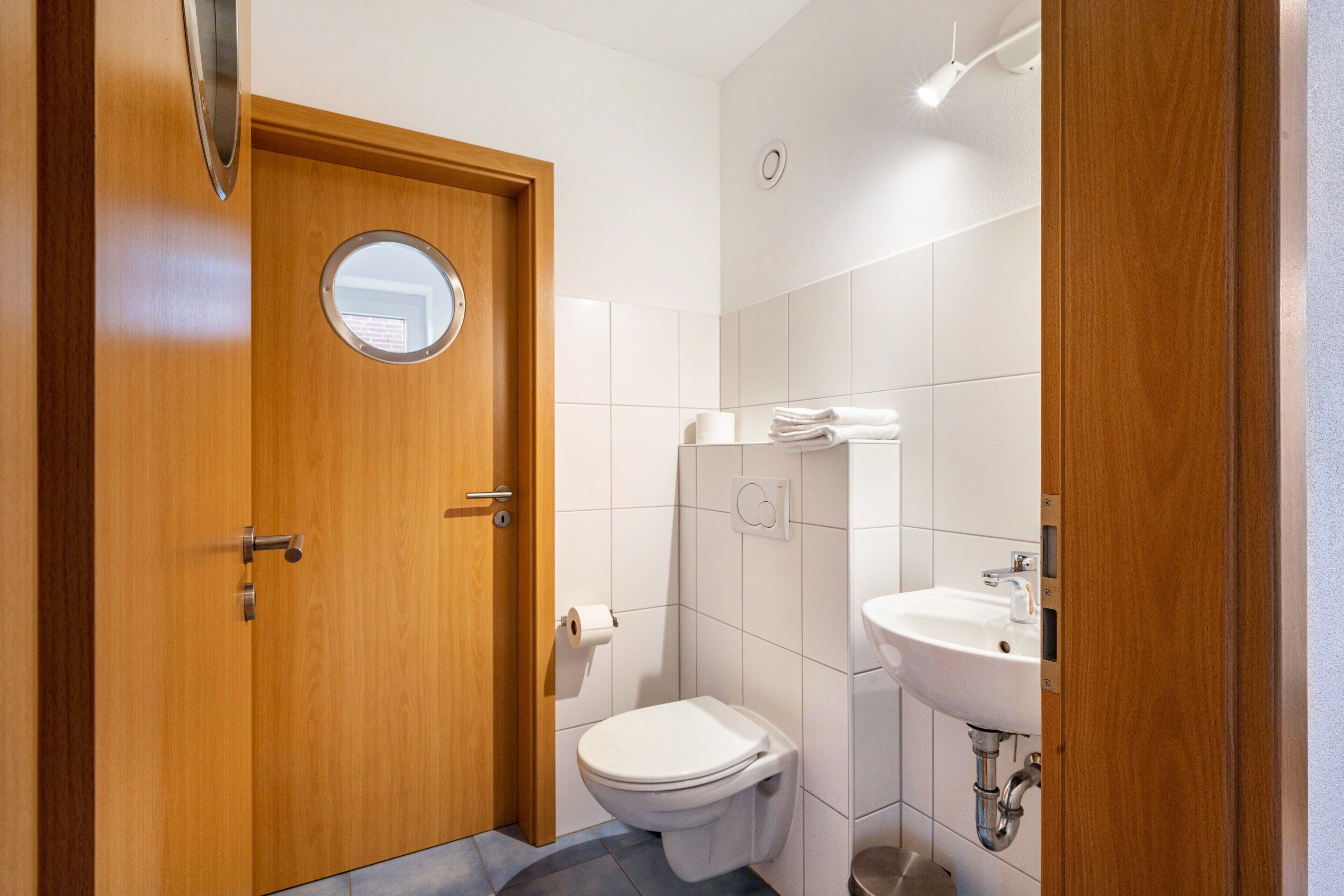 Un bagno piccolo e moderno con una porta in legno, uno specchio rotondo, un water, un lavandino e un porta asciugamani.