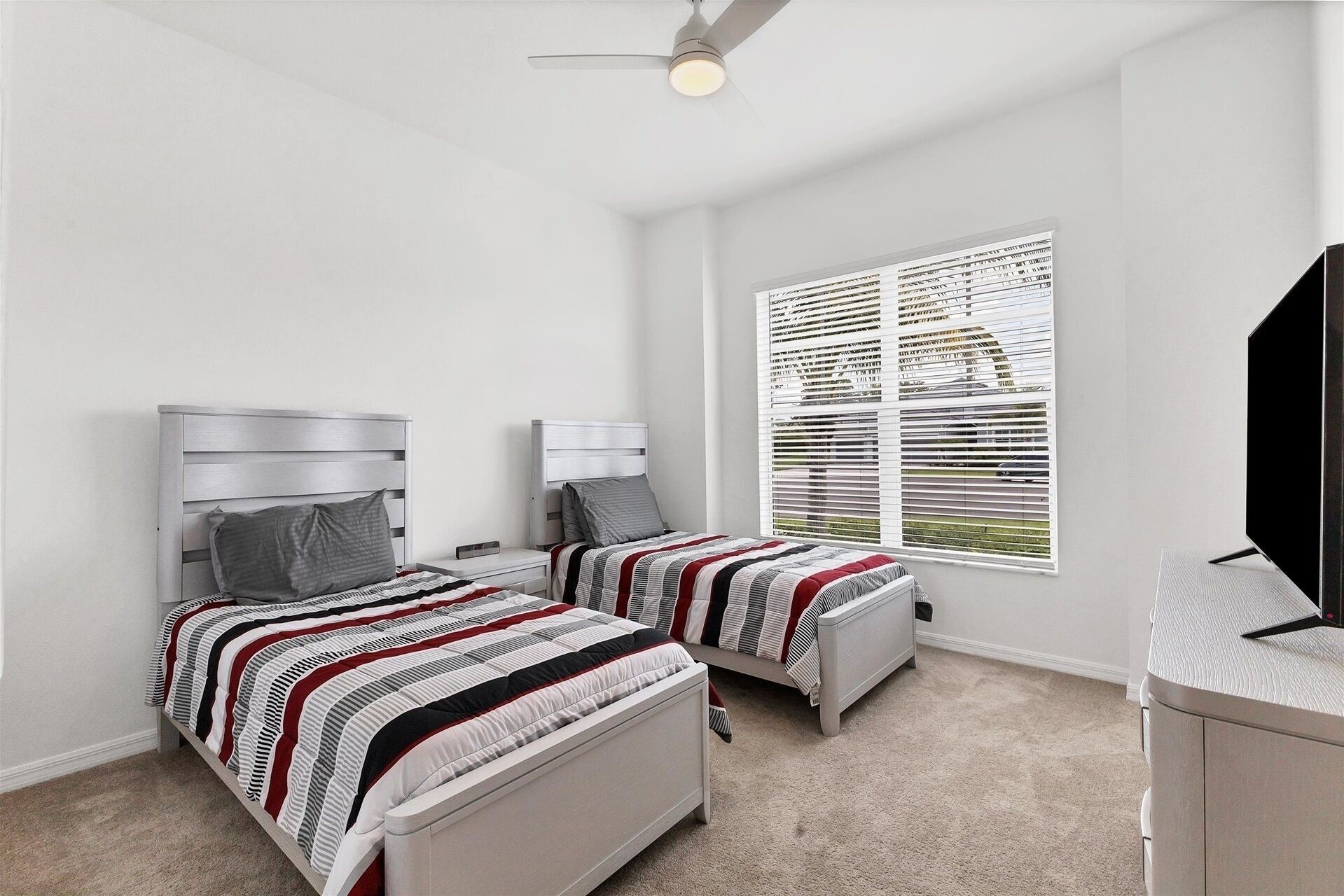 Een slaapkamer met twee bedden, een plafondventilator, een raam met zonwering en een televisie.
