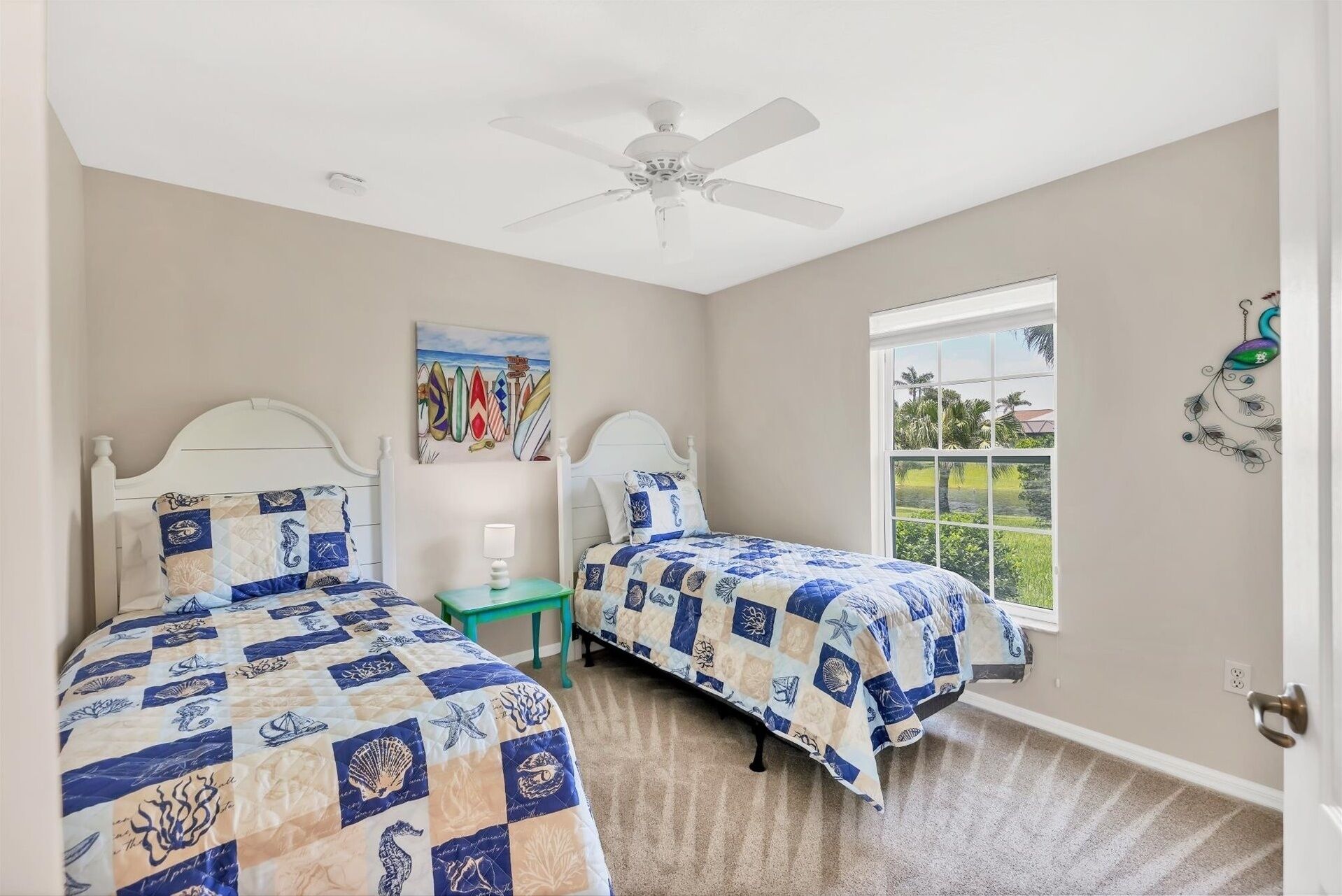 A bedroom with two beds, a ceiling fan, a window with a view of palm trees, and a painting of surfboards.