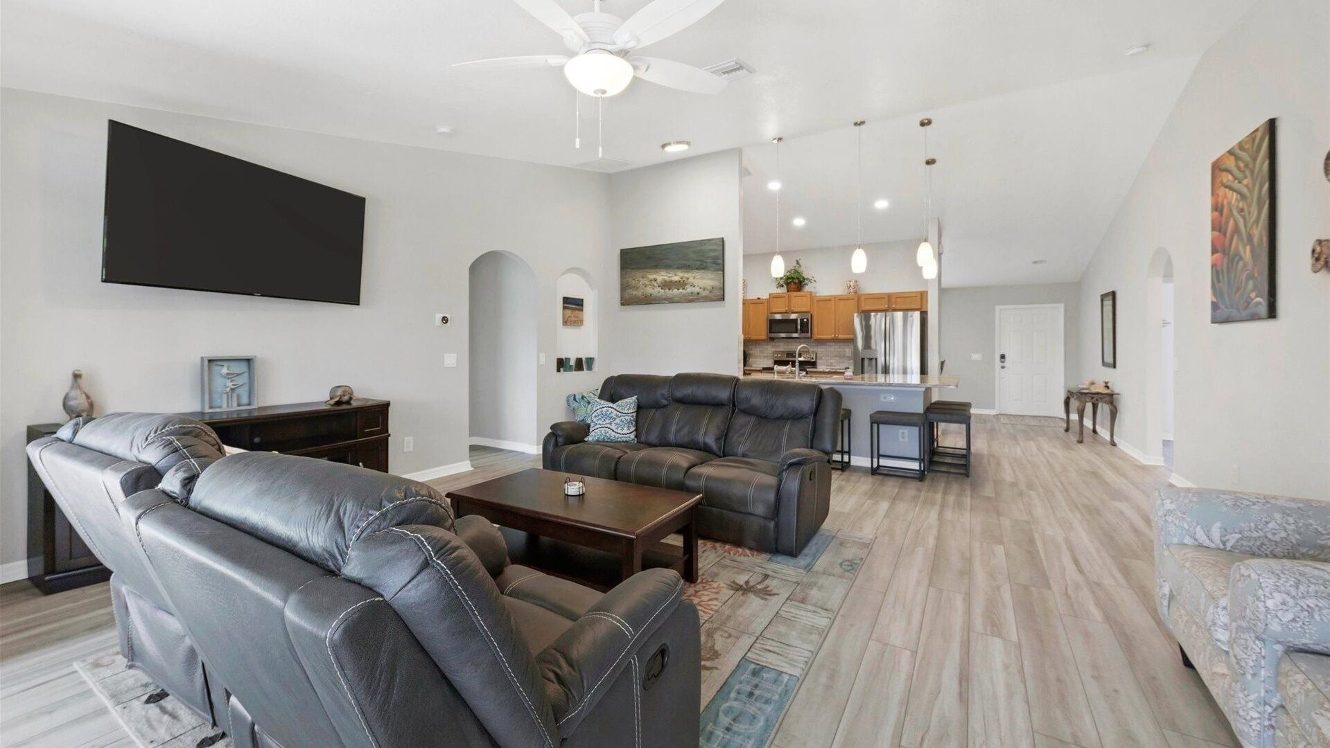 A modern living room with leather sofas, a flat-screen TV, a dining area with a kitchen in the background, and a ceiling fan.