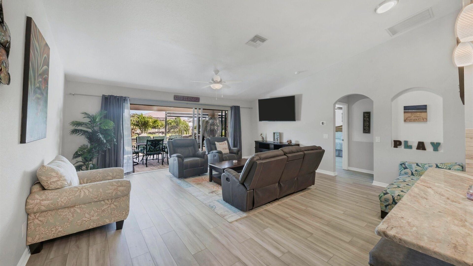 A spacious living area with a sofa, chairs, a coffee table, and a TV. There is a balcony with outdoor seating and a view of palm trees.